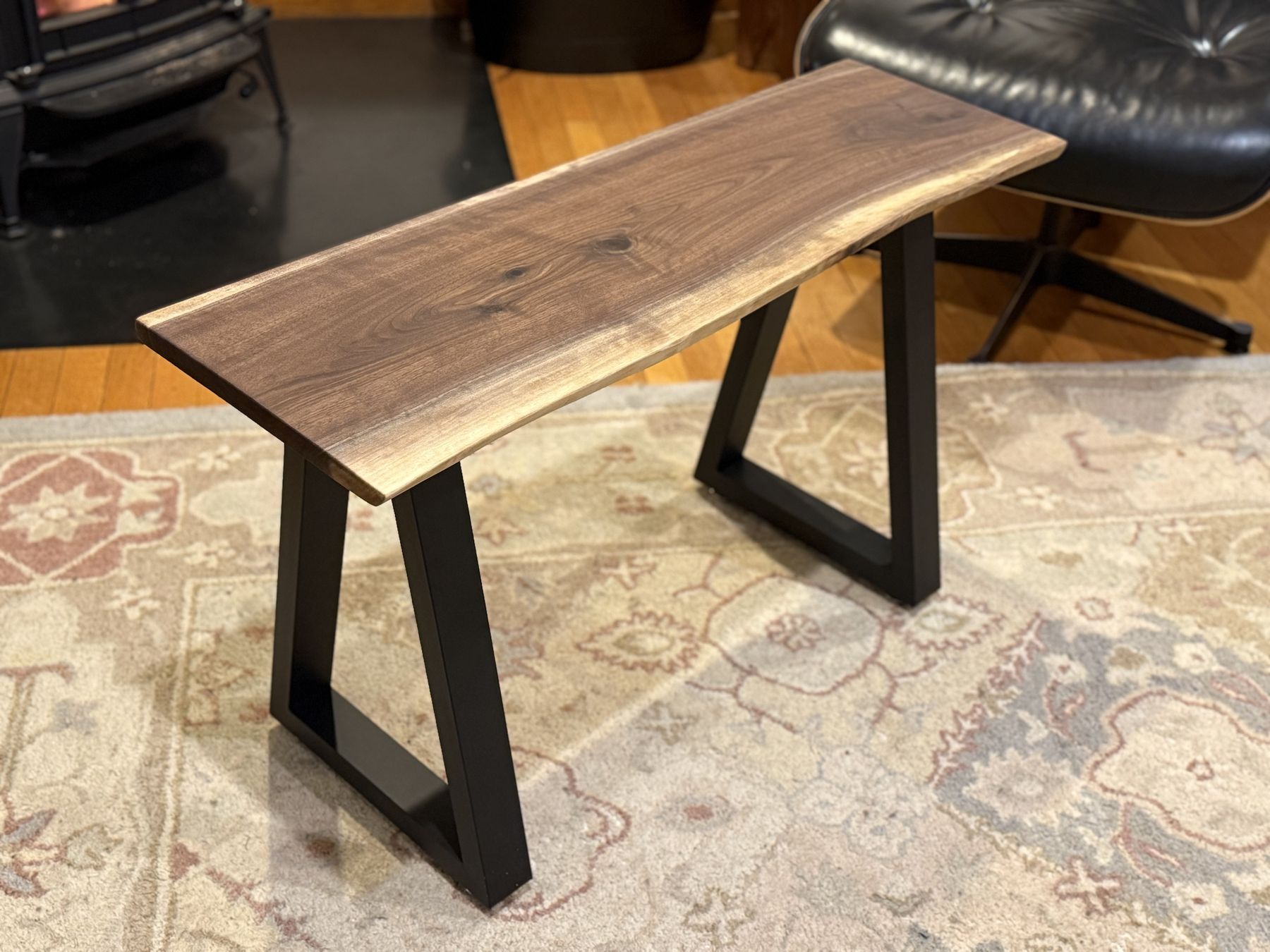 A live-edge walnut bench on black steel trapezoid legs, set on a pale rug beside a leather lounge chair.